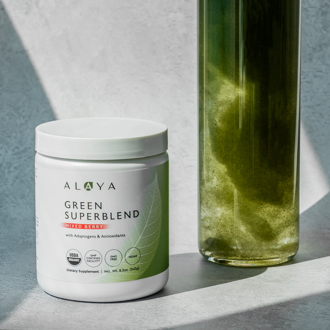 Alaya Green Superblend container with a glass of water and the greens powder on a light gray background