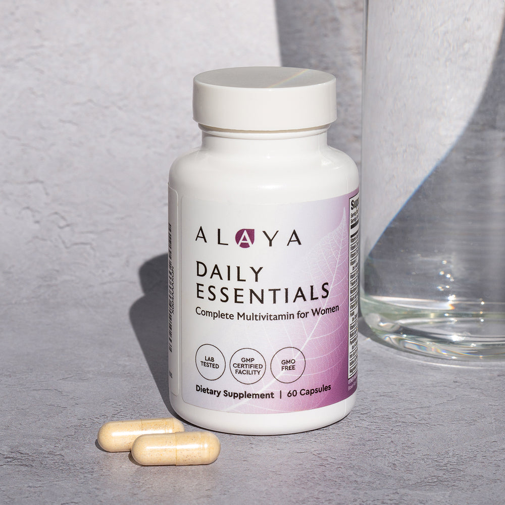 Alaya Daily Essentials multivitamin bottle with two capsules on a gray surface and a glass of water in the background